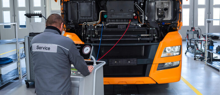 Service technician operating diagnostic equipment in front of an orange truck with its hood open inside a maintenance facility.